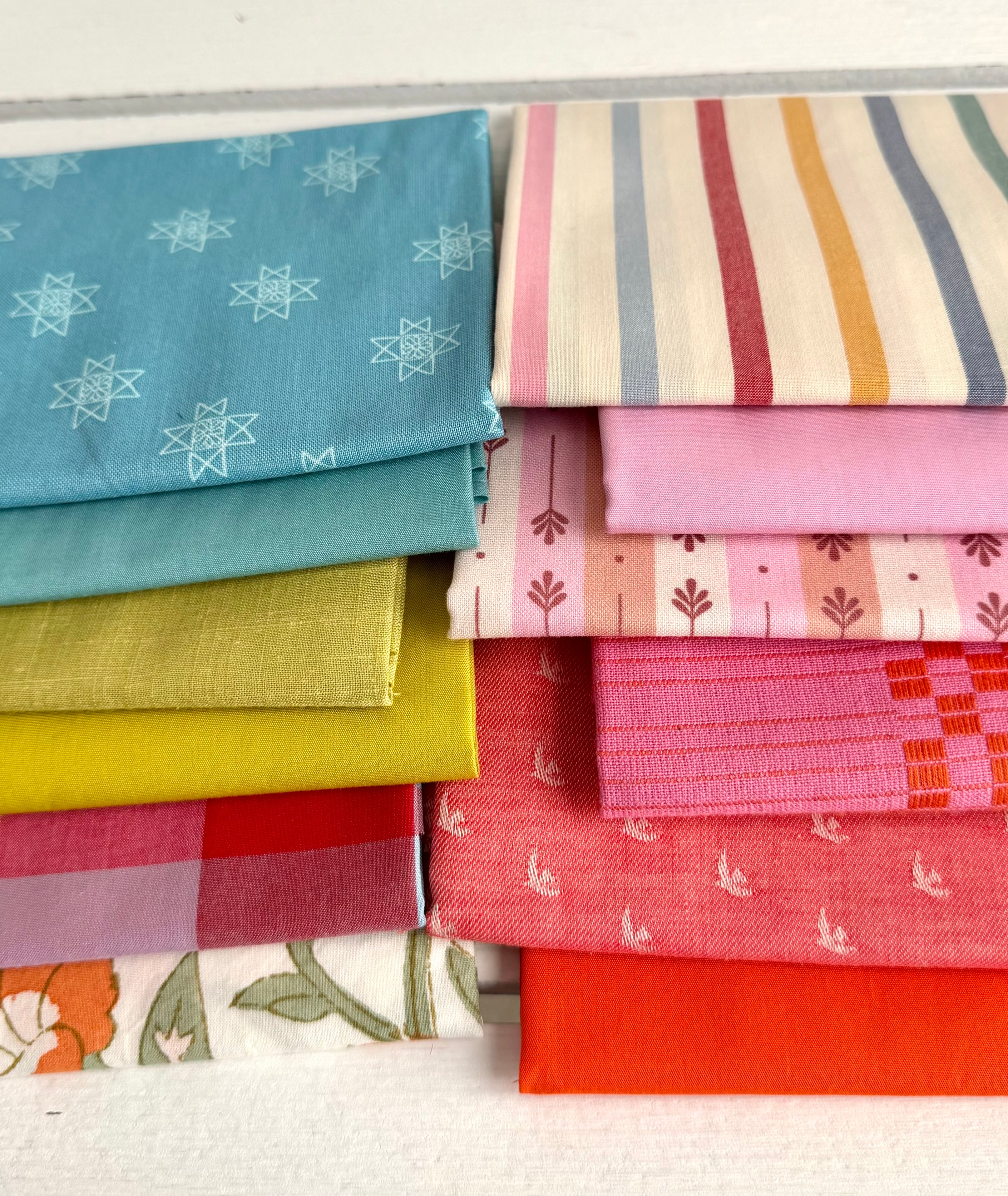 neatly folded set of fabrics stacked against a white wooden background. colors vary from bright orange-red, cream, chartreuse, and warm teals.