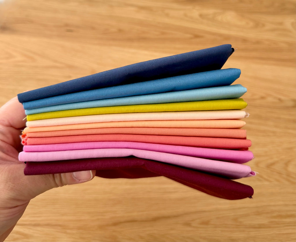 A hand holds a stack of folded fabric swatches in multiple colors, including navy, blue, light blue, yellow, cream, peach, coral, pink, and burgundy, against a light wood background.
