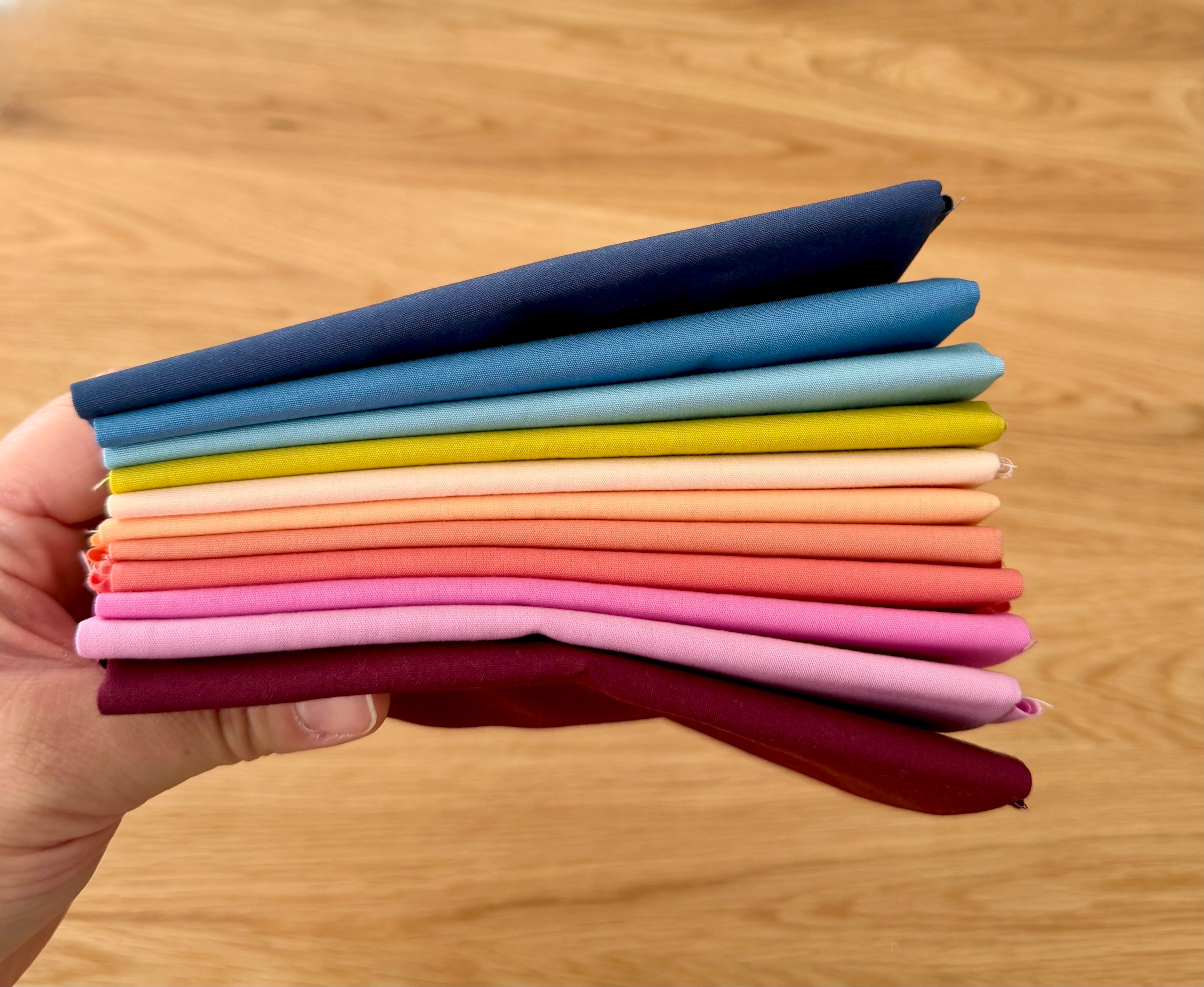 A hand holds a stack of folded fabric swatches in multiple colors, including navy, blue, light blue, yellow, cream, peach, coral, pink, and burgundy, against a light wood background.