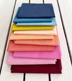 A stack of folded fabric fat quarters in a variety of colors, including navy, blue, light blue, yellow, peach, pink, and burgundy, arranged on a white wooden plank surface.