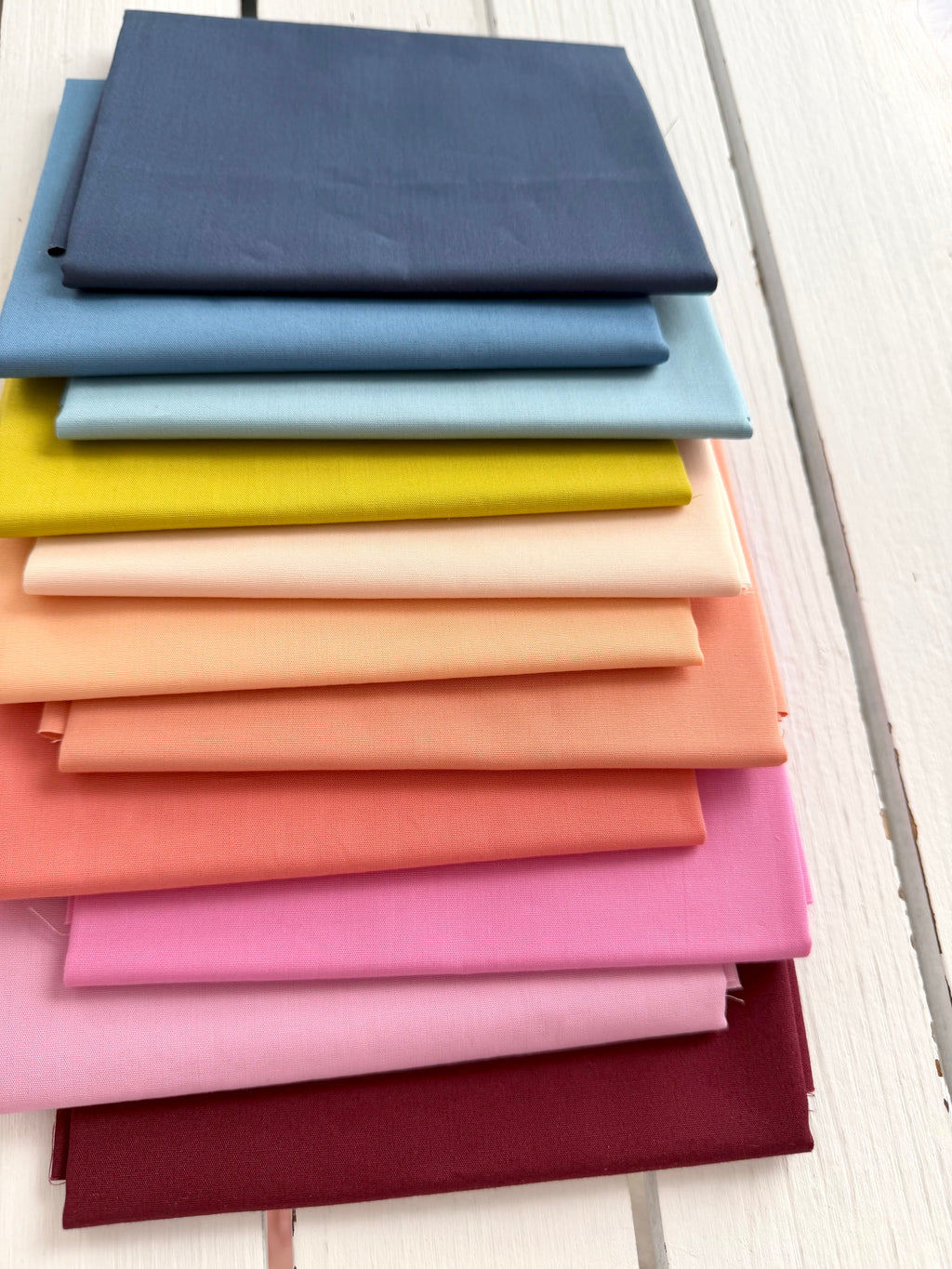 A stack of folded fabric swatches in multiple colors, including navy, light blue, yellow, cream, peach, coral, pink, and burgundy, arranged on a white wooden surface.