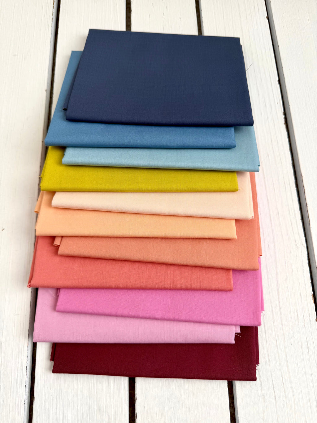 A stack of folded fabric swatches in multiple colors, including navy, blue, yellow, peach, pink, and burgundy, arranged on a white wooden surface.