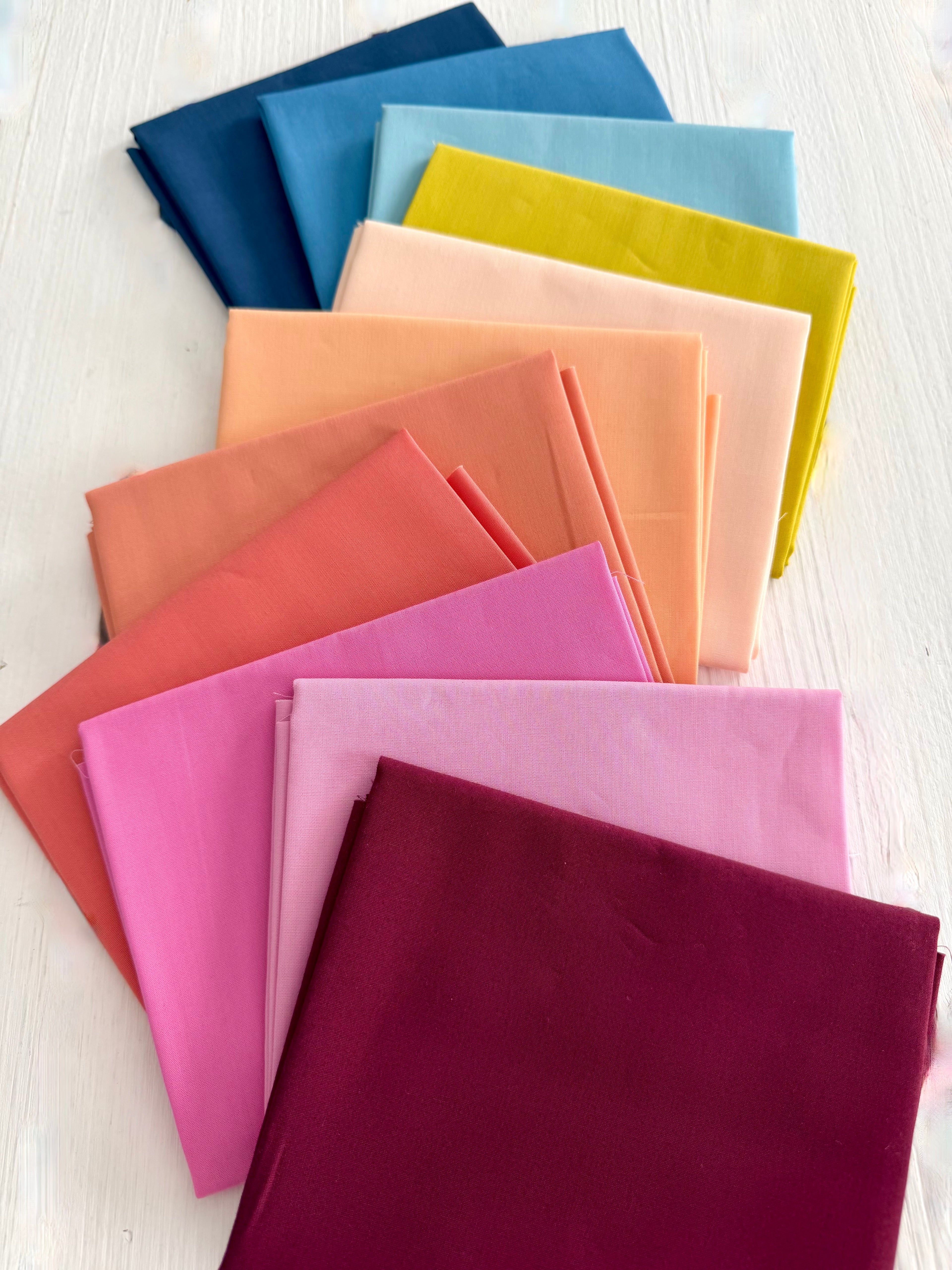 A fan arrangement of folded fabric in multiple colors, including dark slate blue, light blue, chartreuse, peach, coral, pink, and deep burgundy wine, displayed on a white wood surface.