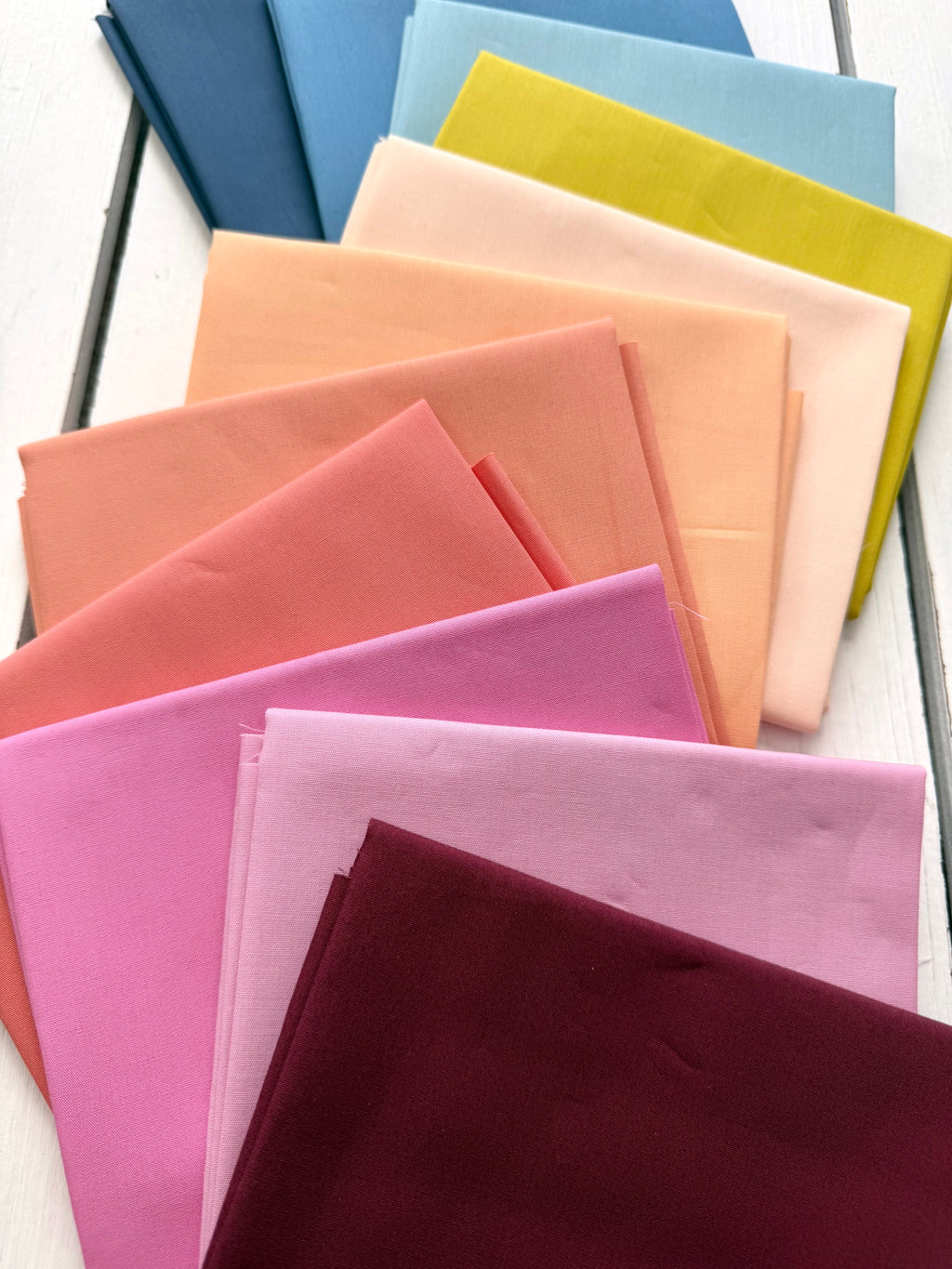 A fan arrangement of folded fabric in multiple pastel and bright colors, including blue, yellow, peach, pink, and burgundy, displayed on a white wood surface.