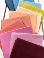 A fan arrangement of folded fabric in multiple pastel and bright colors, including blue, yellow, peach, pink, and burgundy, displayed on a white wood surface.