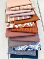 A stack of folded fabric pieces in soft pink, beige, orange, mauve, gray, blue, and white floral/striped patterns, arranged on a white wood surface.