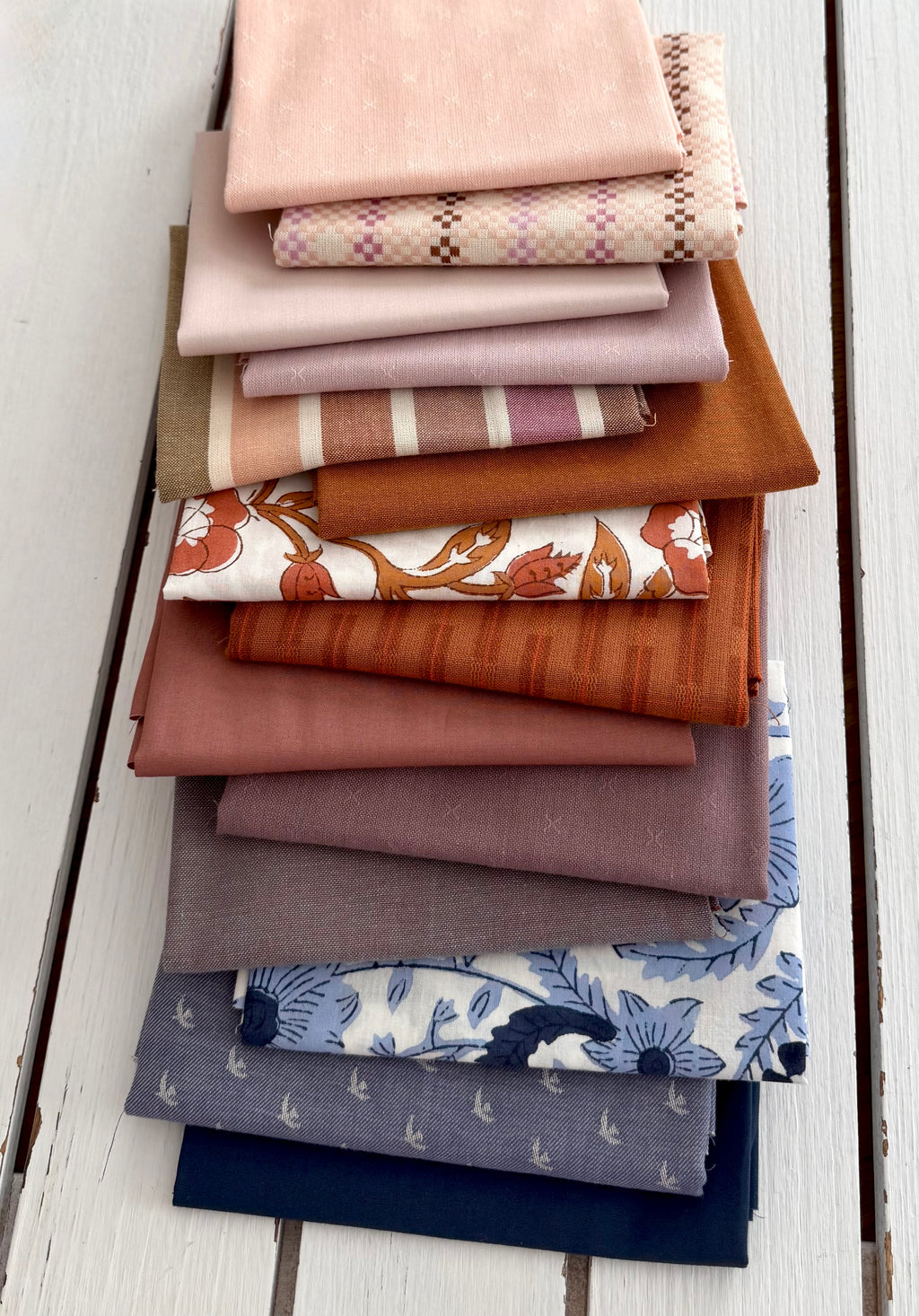 A stack of folded fabric swatches in various colors and patterns, including solid blush, plaid, stripes, rust orange, floral, and blue botanical prints, arranged on a white wooden plank surface.