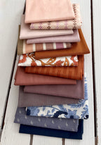 A stack of folded fabric swatches in various colors and patterns, including solid blush, plaid, stripes, rust orange, floral, and blue botanical prints, arranged on a white wooden plank surface.