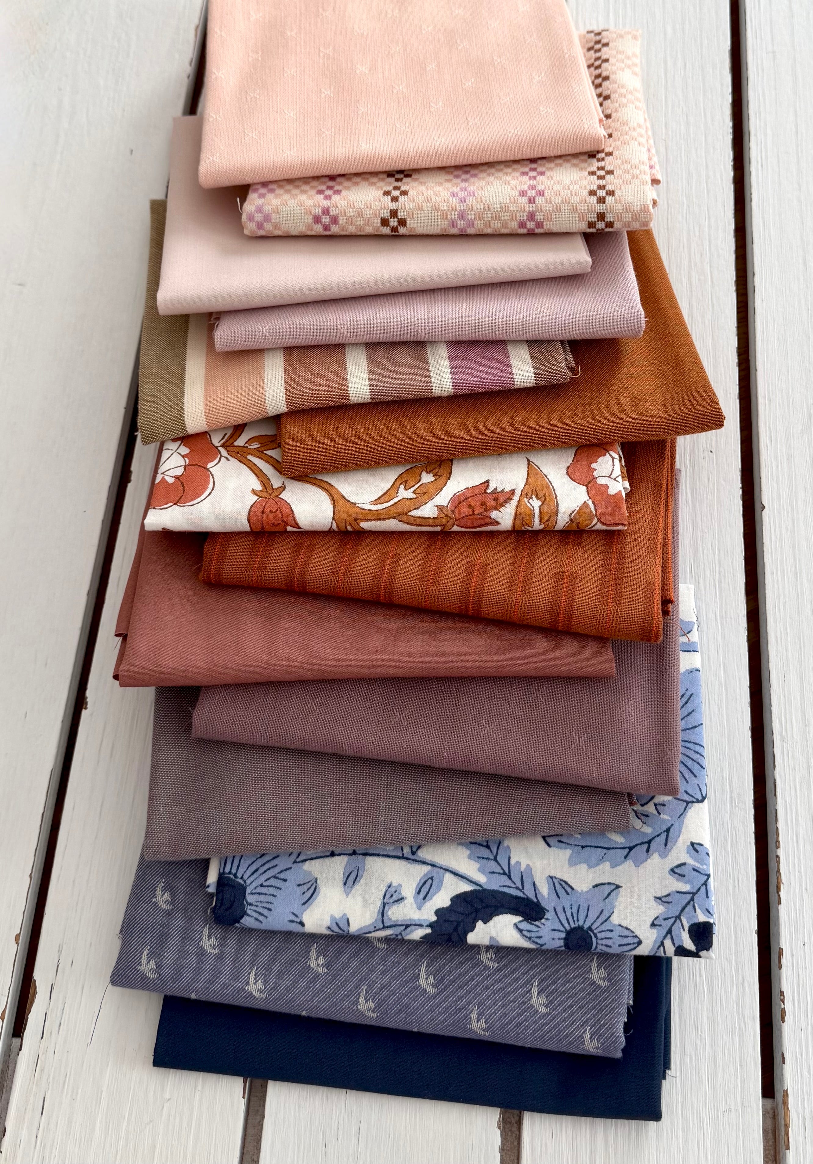 A stack of folded fabric swatches in various colors and patterns, including solid blush, plaid, stripes, rust orange, floral, and blue botanical prints, arranged on a white wooden plank surface.