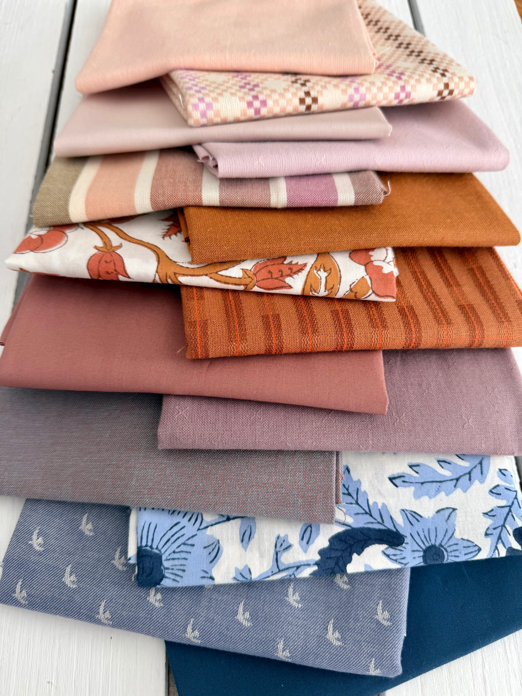 A stack of folded fabric pieces in assorted colors and patterns, including solid blush, pink, orange, blue, and floral prints, arranged on a white wooden surface.