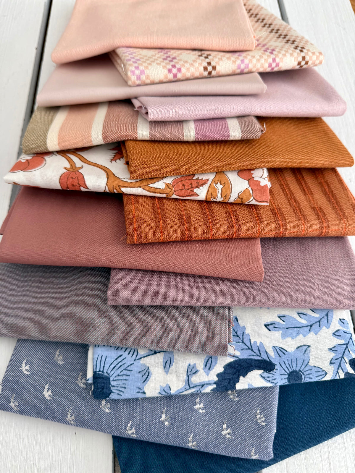 A stack of folded fabric pieces in assorted colors and patterns, including solid blush, pink, orange, blue, and floral prints, arranged on a white wooden surface.