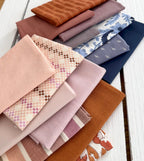 A stack of folded fabric samples in soft pastel and earthy tones, including blush pink, dusty rose, mauve, navy, cream, and orange-brown, arranged on a white painted wooden surface.
