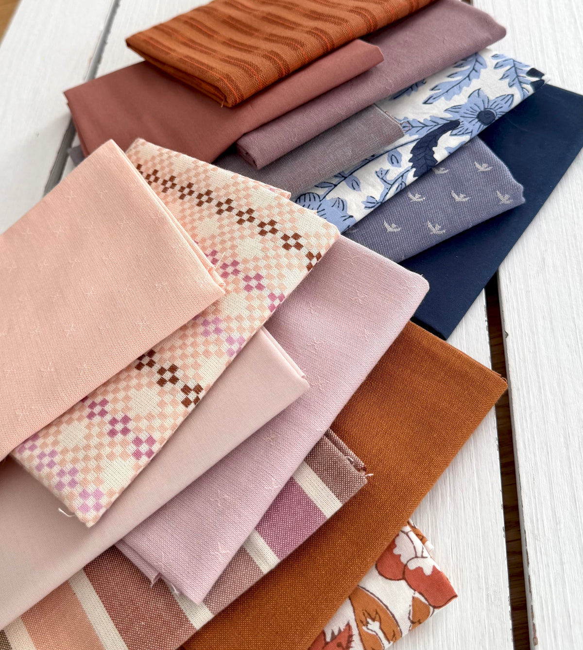 A stack of folded fabric samples in soft pastel and earthy tones, including blush pink, dusty rose, mauve, navy, cream, and orange-brown, arranged on a white painted wooden surface.