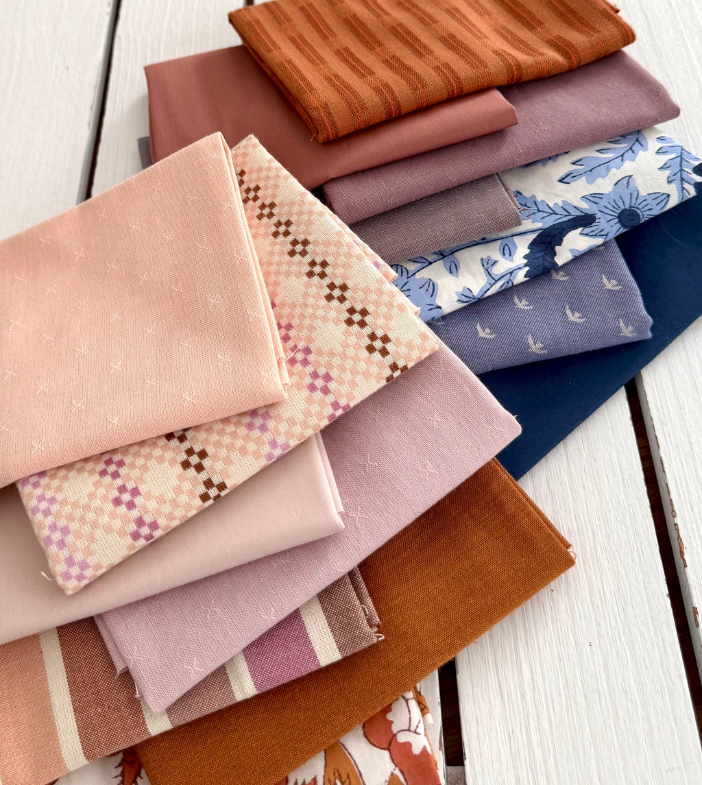 A stack of folded fabric pieces in soft pink, dusty rose, rust orange, blue floral, and navy tones, arranged on a white wooden surface.