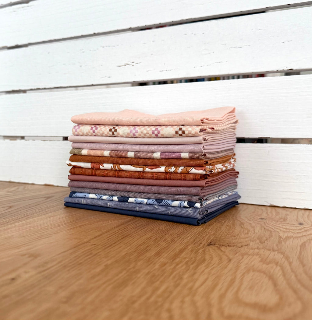 A stack of folded patterned fabrics in soft pink, beige, brown, orange, blue, and white, arranged on a wooden surface in front of a white wooden slat wall.