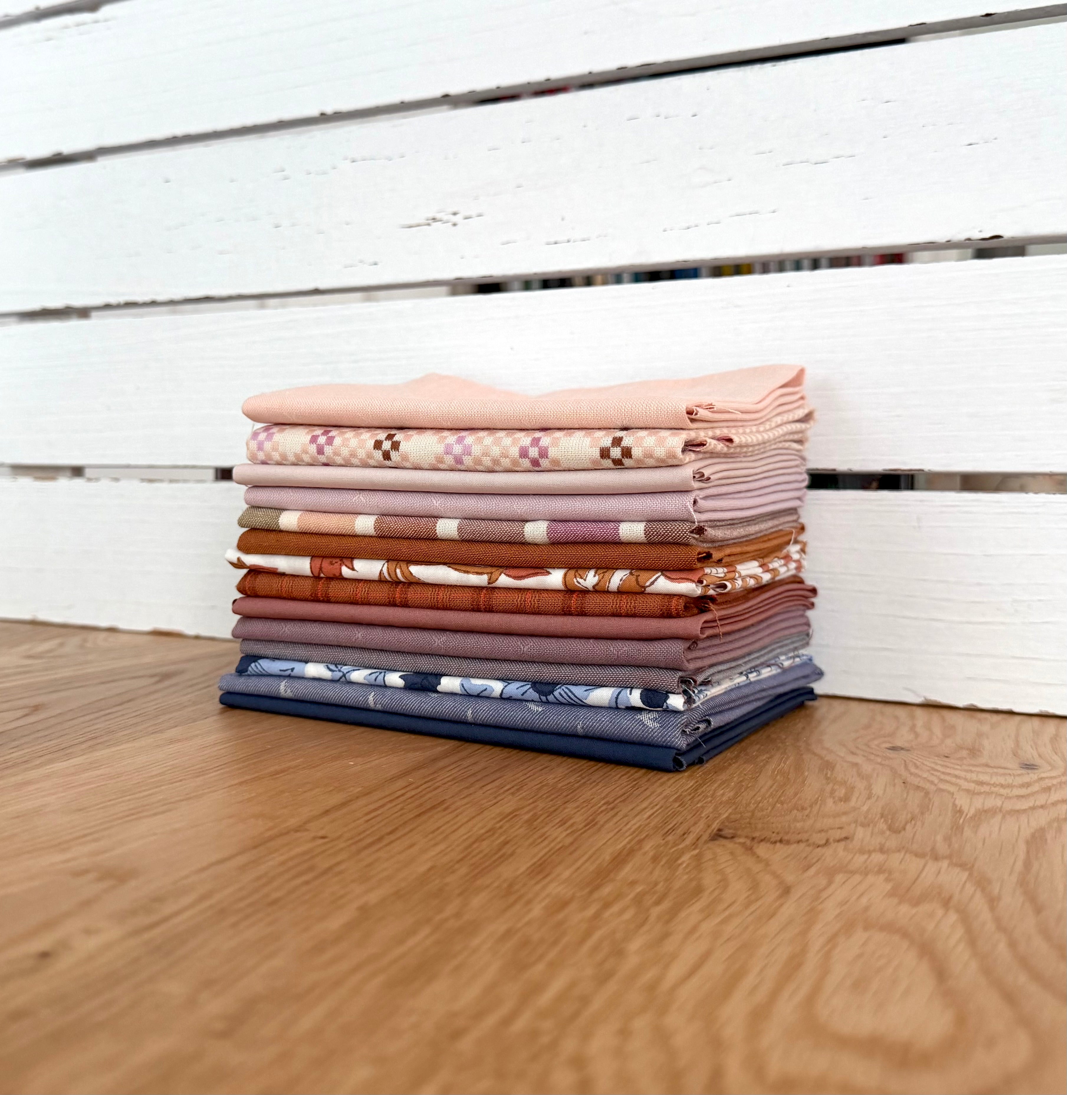 A stack of folded patterned fabrics in soft pink, beige, brown, orange, blue, and white, arranged on a wooden surface in front of a white wooden slat wall.