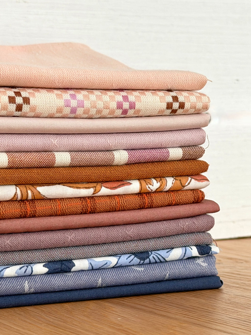A stack of folded patterned fabrics in soft neutral and earthy tones, including blush, beige, pink, brown, orange, mauve, gray, and navy, arranged on a wooden surface against a light wall.
