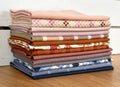A stack of folded fabric pieces in assorted colors and patterns, including solid blush, pink-and-white check, tan, rust, cream, and navy floral prints, arranged on a wooden surface against a white paneled background.