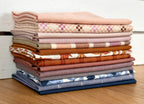 A stack of folded fabric pieces in assorted colors and patterns, including solid blush, pink-and-white check, tan, rust, cream, and navy floral prints, arranged on a wooden surface against a white paneled background.