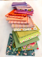 A stack of folded fabric pieces in assorted colors and patterns, including purple, orange, pink, peach, cream, green, and floral prints, arranged on a white wooden surface.