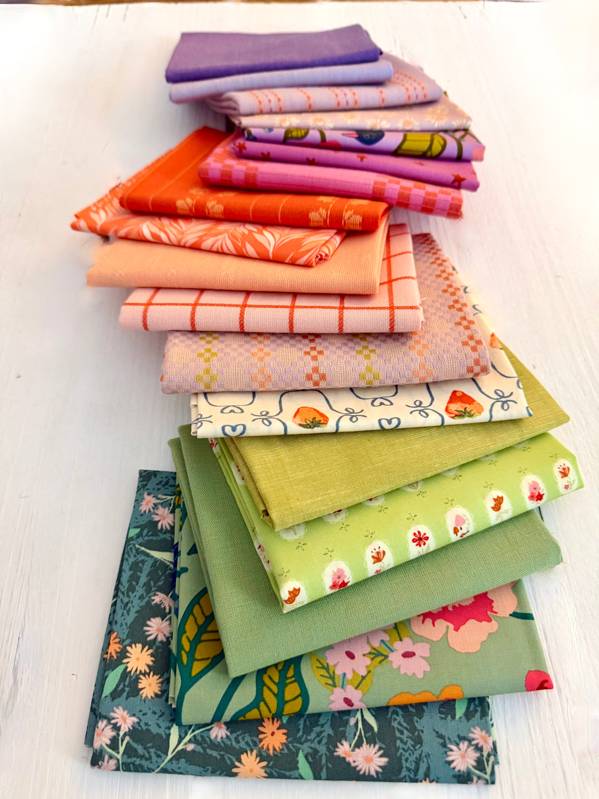A stack of folded fabric pieces in assorted colors and patterns, including purple, orange, pink, peach, cream, green, and floral prints, arranged on a white wooden surface.