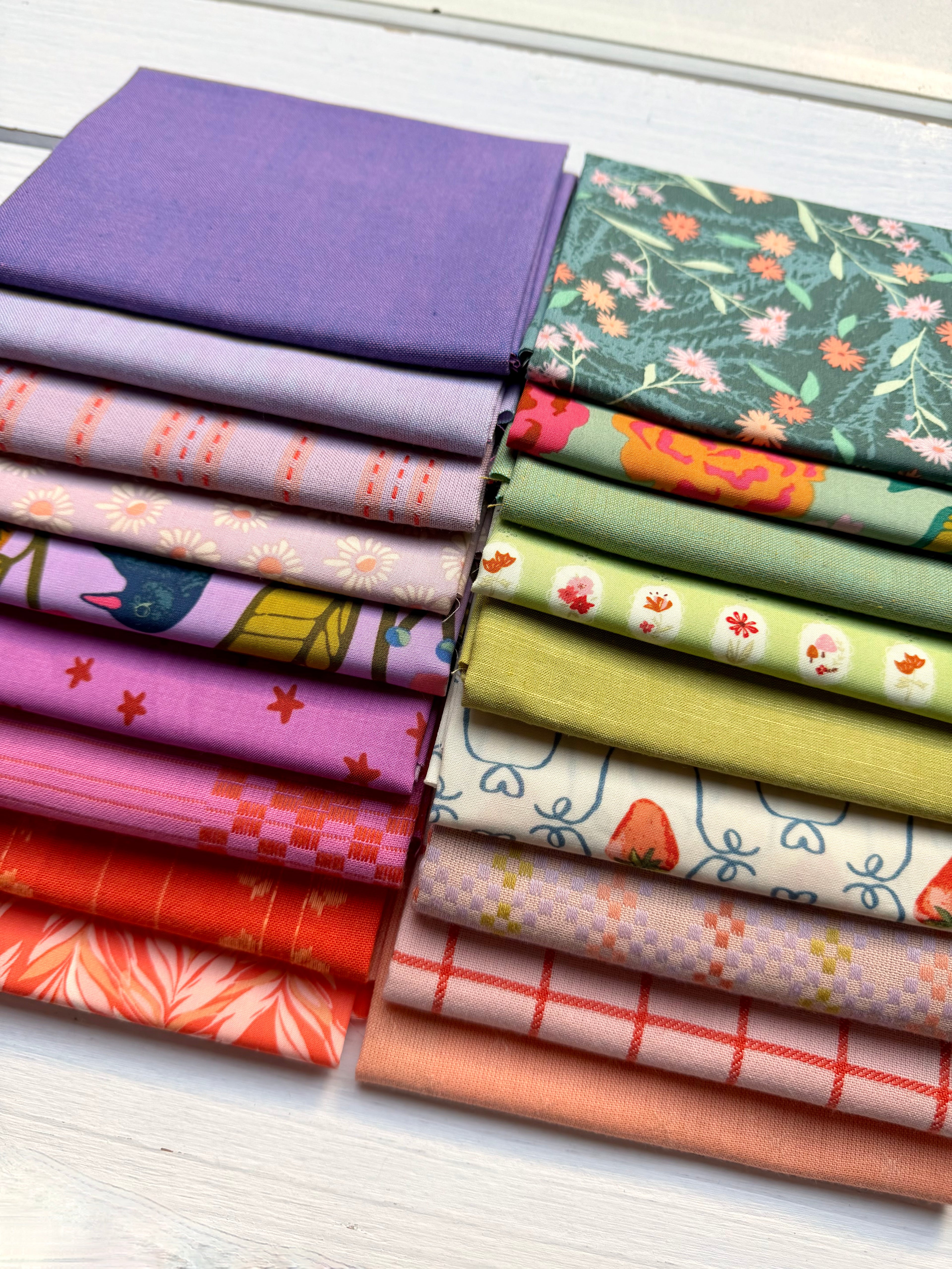 Two stacks of folded fabric samples in assorted colors and patterns, including solid purple, floral green, pink, orange, red, and plaid designs, arranged on a white wood surface.