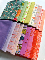 A set of folded fabric pieces in assorted colors and patterns, including floral prints, stripes, checks, and solid colors, arranged diagonally on a white textured surface.