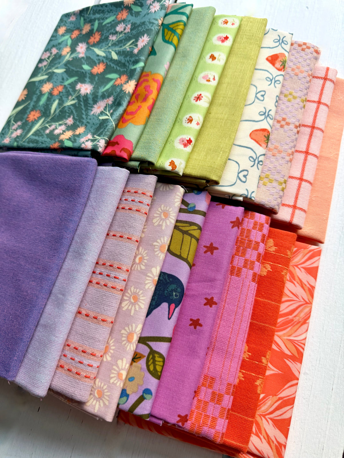 A set of folded fabric pieces in assorted colors and patterns, including floral prints, stripes, checks, and solid colors, arranged diagonally on a white textured surface.