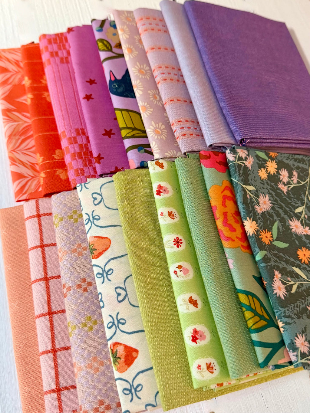 A collection of folded fabric swatches in various colors and patterns, including solid purple, orange, pink, green, and floral prints, arranged neatly on a white wooden surface.
