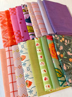 A collection of folded fabric swatches in various colors and patterns, including solid purple, orange, pink, green, and floral prints, arranged neatly on a white wooden surface.