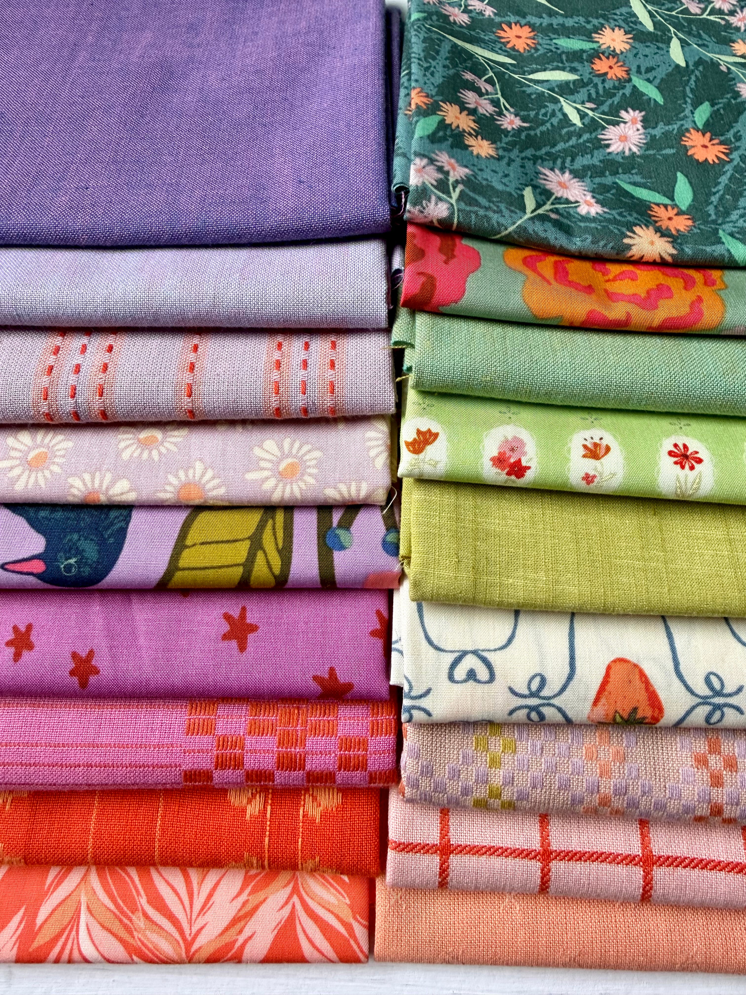 A stack of folded fabric pieces in multiple colors and patterns, including solid purple, floral green, orange and pink florals, blue and green bird motifs, white with red flowers, red plaid, and peach plaid.