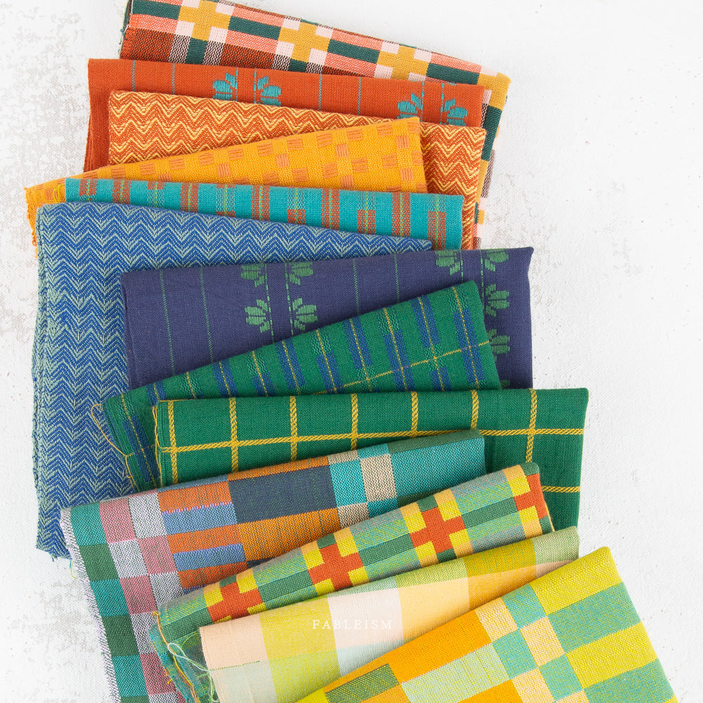 A stack of colorful patterned fabric squares in various shades of blue, green, orange, yellow, and plaid, arranged diagonally on a light textured background.